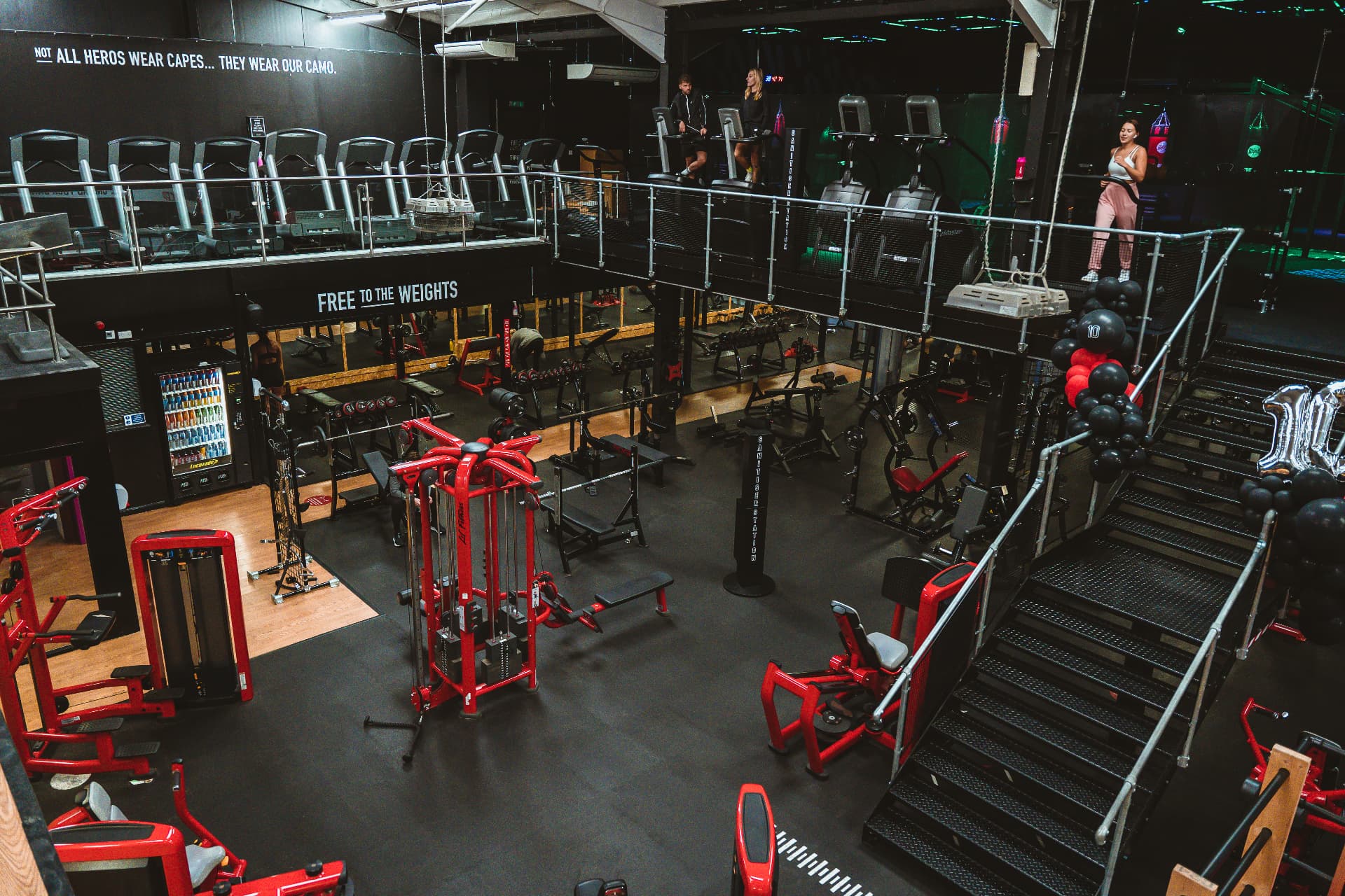 Wide view of the Heros floor: red Life Fitness machines below, cardio deck above the 'Free to the Weights' wall